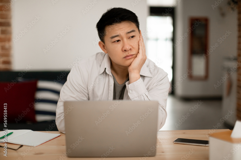 Upset bored middle aged asian man sitting in front of laptop at home, leaning on his hand and looking away Upset bored middle aged asian man sitting in front of laptop at home, leaning on his hand and looking away