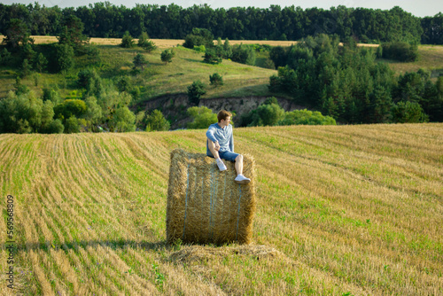 Young man in blue clothes is sitting on a bale of straw on the background of a field
