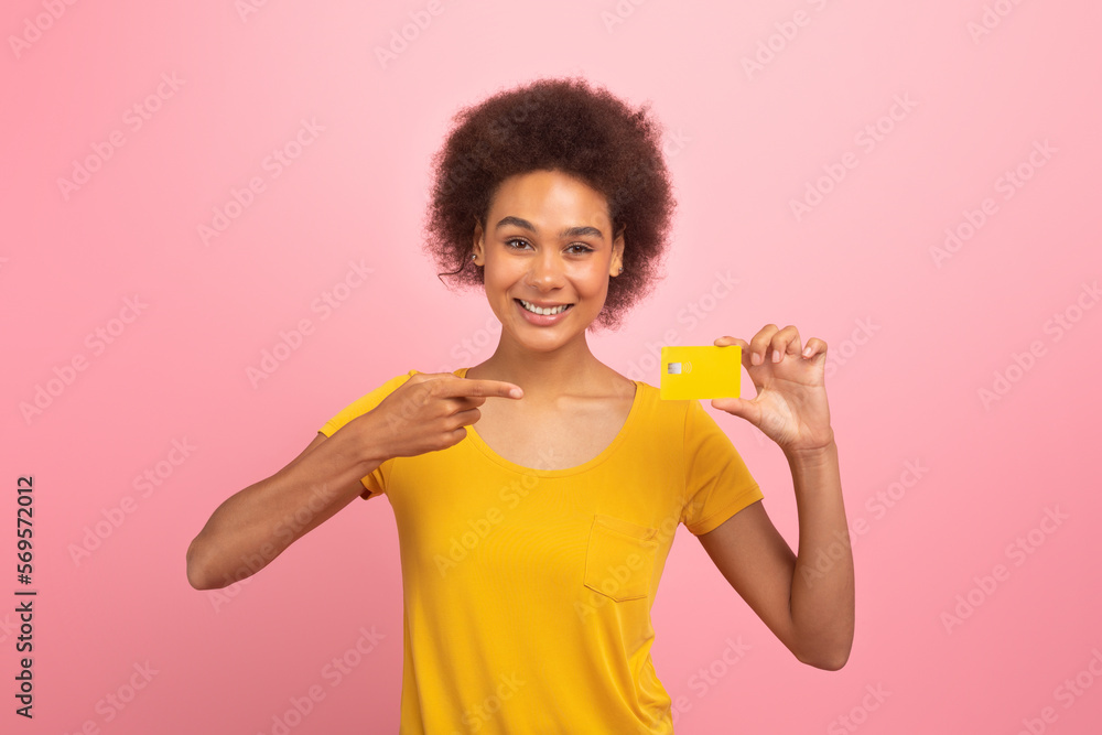 Cheerful young african american curly lady shopaholic in yellow t-shirt point finger at credit card