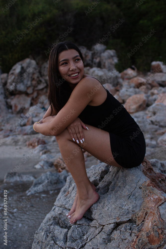 Beautiful woman in black dress smiling while sitting on the beach..A cheerful woman in a black dress sits on a rocky mountain and looks away, relaxing near the sea in summer. Beautiful woman in black dress smiling while sitting on the beach..A cheerful woman in a black dress sits on a rocky mountain and looks away, relaxing near the sea in summer.