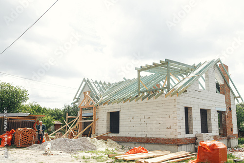 Unfinished modern farmhouse building. Wooden roof framing of mansard with dormer and aerated concrete block walls. Timber trusses, rafters and beams on blocks. New house construction photo