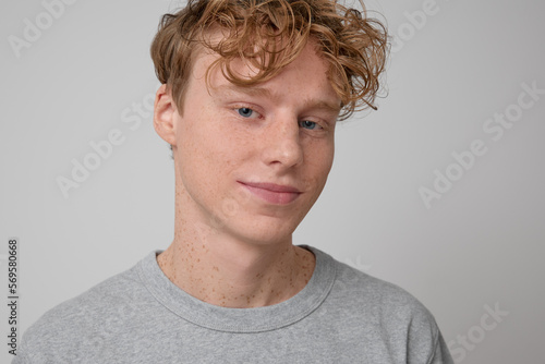 Close up of happy handsome freckled red haired smiling teenager with blue eyes wearing gray t-shirt casual wear showing emotions and looking at the camera posing for photo against the gray backdrop. 