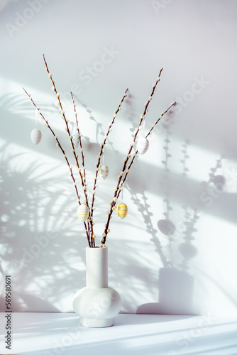 Easter home decor. Modern minimalist style interior with blooming branches of the pussy willow and hanging easter eggs in ceramic vase on a white console with sunlight, shadows on gray wall. Vertical.