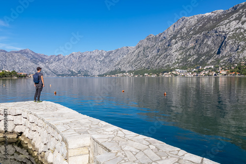 Hiking man in small port in Kotor Bay in coastal town Prcanj, Adriatic Mediterranean Sea, Montenegro, Balkans, Europe. Scenic view on village Dobrota. Fjord winding along beautiful Dinaric Alps