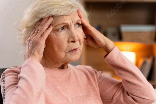 Portrait of attractive senior woman sitting on sofa at home with headache, feeling pain and with expression of being unwell. Heath problems concept