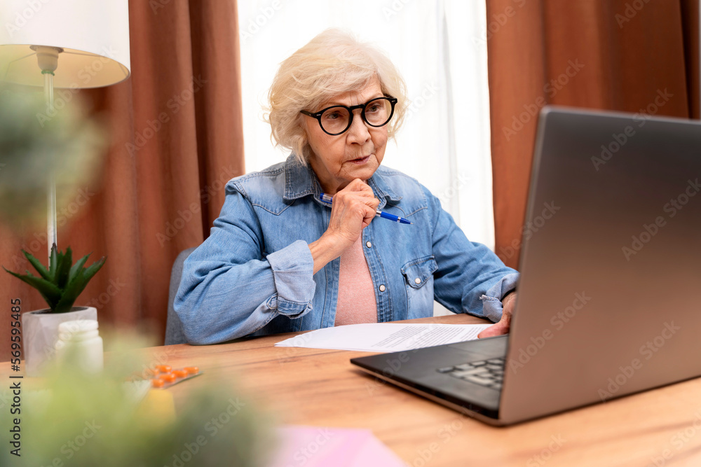 Serious senior woman holding online training while working at home with laptop. Old woman wearing eyeglasses teaching students online. Beautiful stylish elderly lady browsing site on computer Serious senior woman holding online training while working at home with laptop. Old woman wearing eyeglasses teaching students online. Beautiful stylish elderly lady browsing site on computer