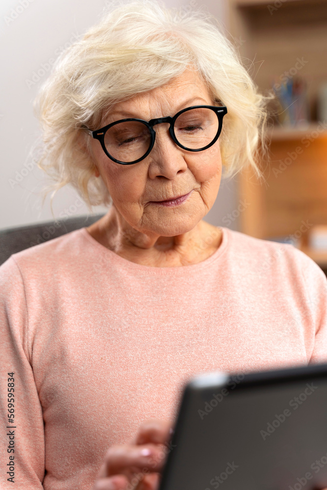 Vertical view of the smiling senior woman sitting with tablet on couch at home and hanging on social media on sunday afternoon. Elderly people and technologies concept Vertical view of the smiling senior woman sitting with tablet on couch at home and hanging on social media on sunday afternoon. Elderly people and technologies concept