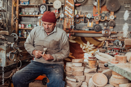 A senior man sitting in the workshop and processing wooden utensils in the old manual way