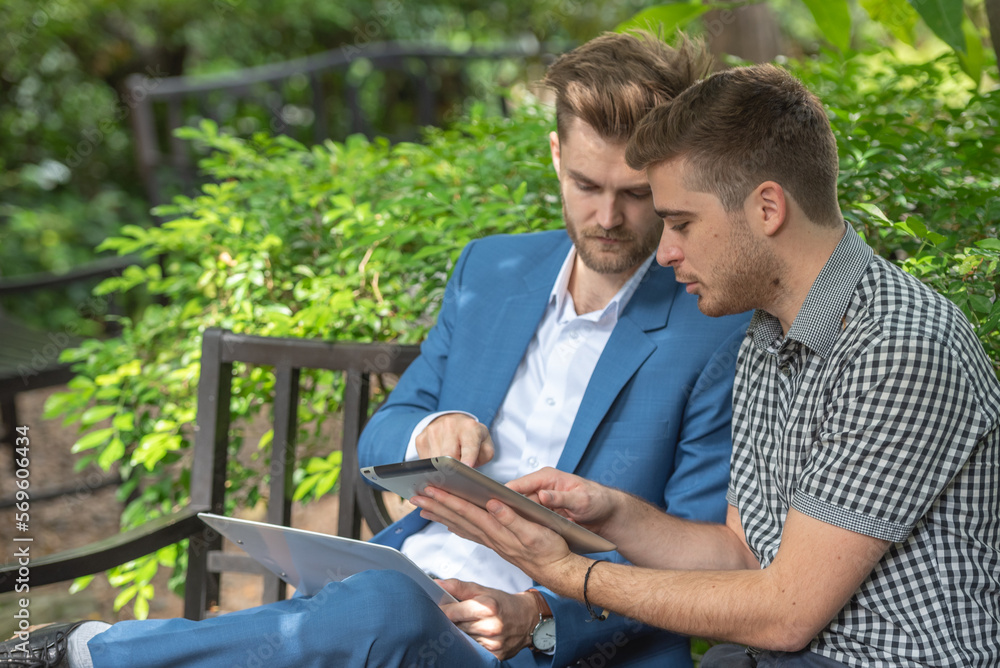 Two businessman or gay sitting at chair in park while using digital tablet and paper work plan for talk discussion team working online togetherness while consulting. Two businessman or gay sitting at chair in park while using digital tablet and paper work plan for talk discussion team working online togetherness while consulting.