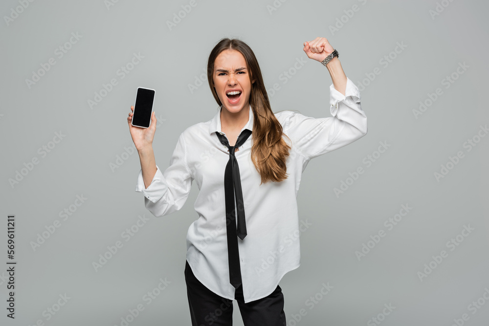 emotional woman in shirt and tie standing with clenched fist and holding smartphone with blank screen isolated on grey, gender equality. emotional woman in shirt and tie standing with clenched fist and holding smartphone with blank screen isolated on grey, gender equality.
