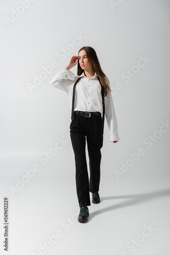full length of brunette young woman in black pants and suspenders walking on grey.