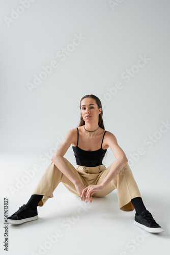 full length of pretty model in beige pants and black top looking at camera while sitting on grey.