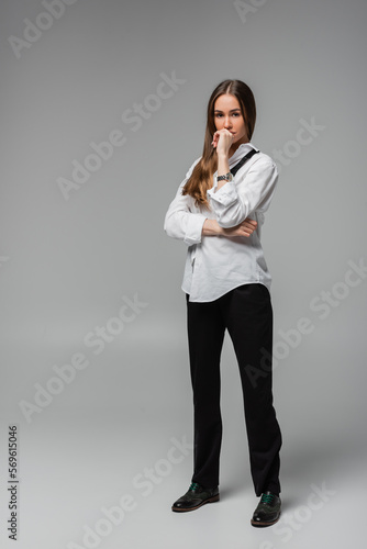 full length of pensive woman in shirt with tie and black pants standing on grey.
