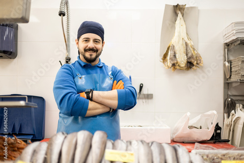 A smiling middle-aged fishmonger poses with his arms crossed behind the sales counter, selling food, small and medium business concept.