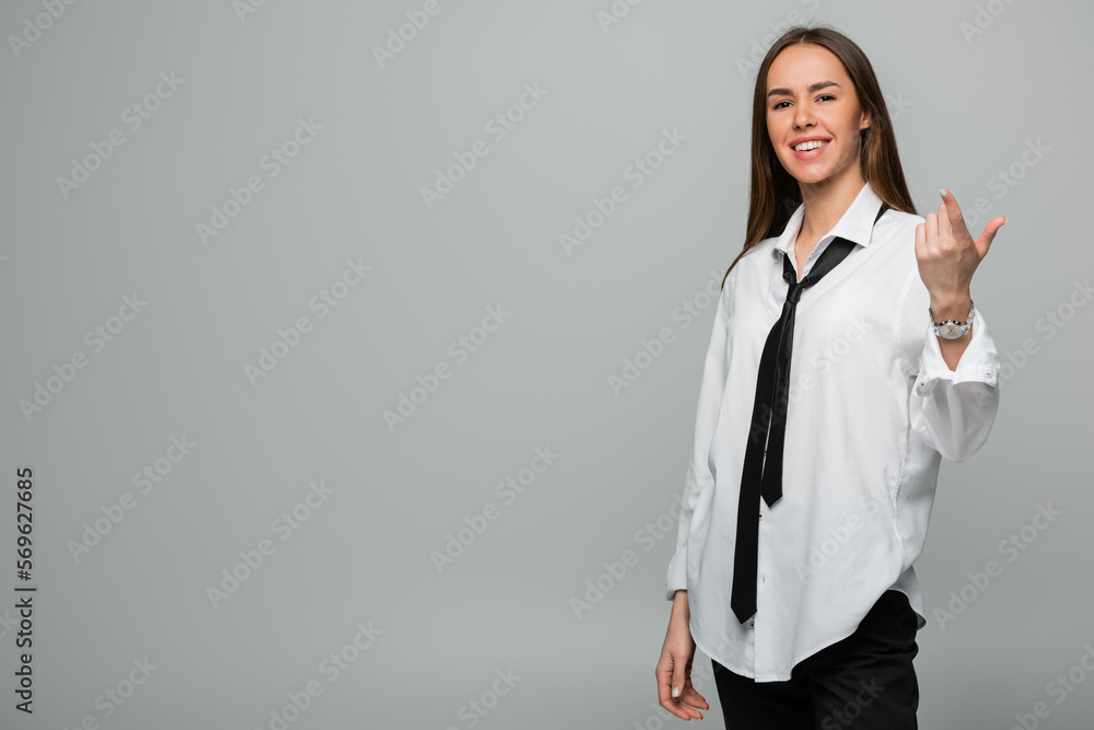 cheerful young woman in white shirt and tie looking at camera while pointing with finger isolated on grey. cheerful young woman in white shirt and tie looking at camera while pointing with finger isolated on grey.