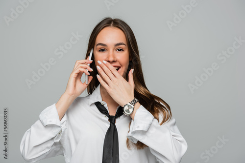happy woman in white shirt with tie talking on smartphone and smiling while covering mouth isolated on grey.