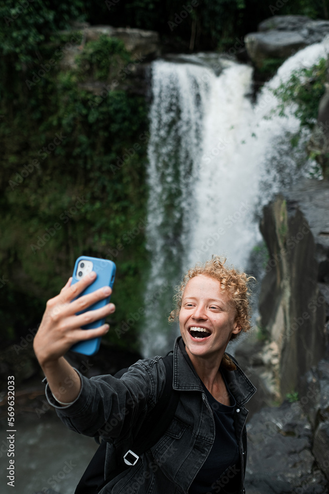 Fototapeta premium A young beautiful and cheerful girl takes a selfie on the phone against the backdrop of a waterfall.