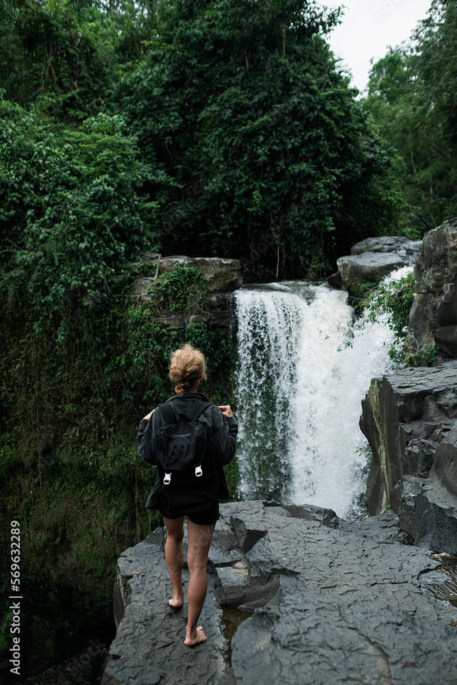 A young blonde girl with a backpack travels to a large waterfall, rear view. A young blonde girl with a backpack travels to a large waterfall, rear view.