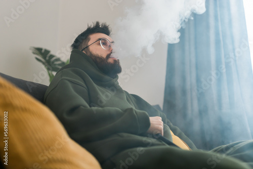 Bearded man is smoking hookah at home and blowing cloud of smoke - chill time and resting concept