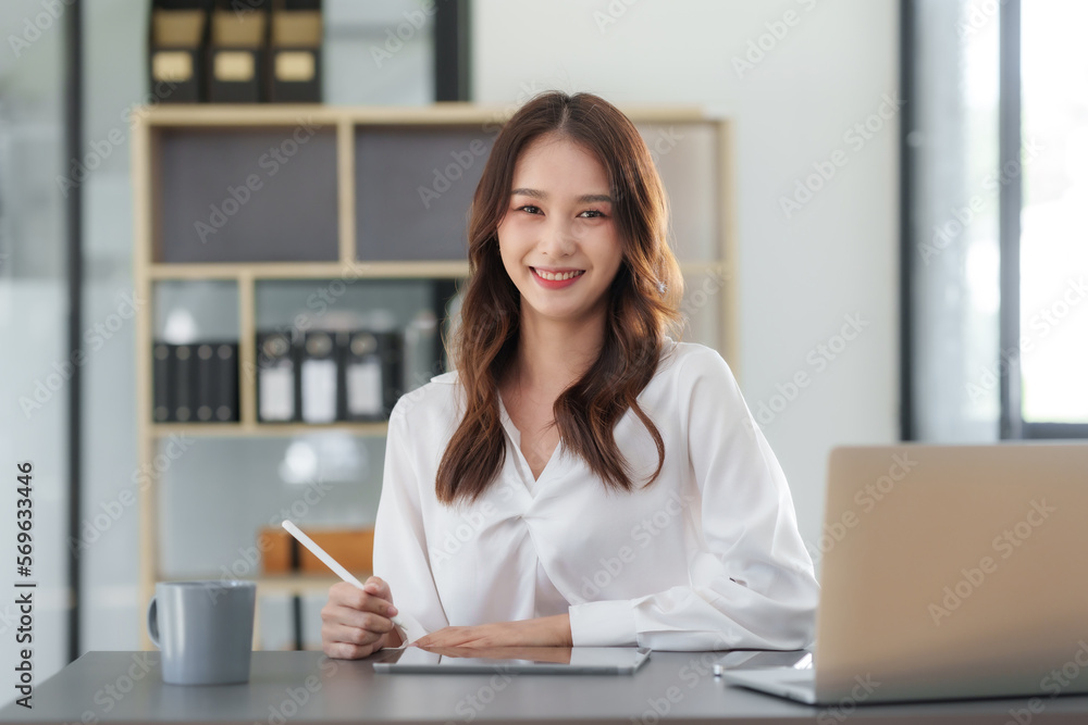 Pretty Asian businesswoman sitting smiling and happy working on laptop computer and document in office. Looking at camera. Pretty Asian businesswoman sitting smiling and happy working on laptop computer and document in office. Looking at camera.