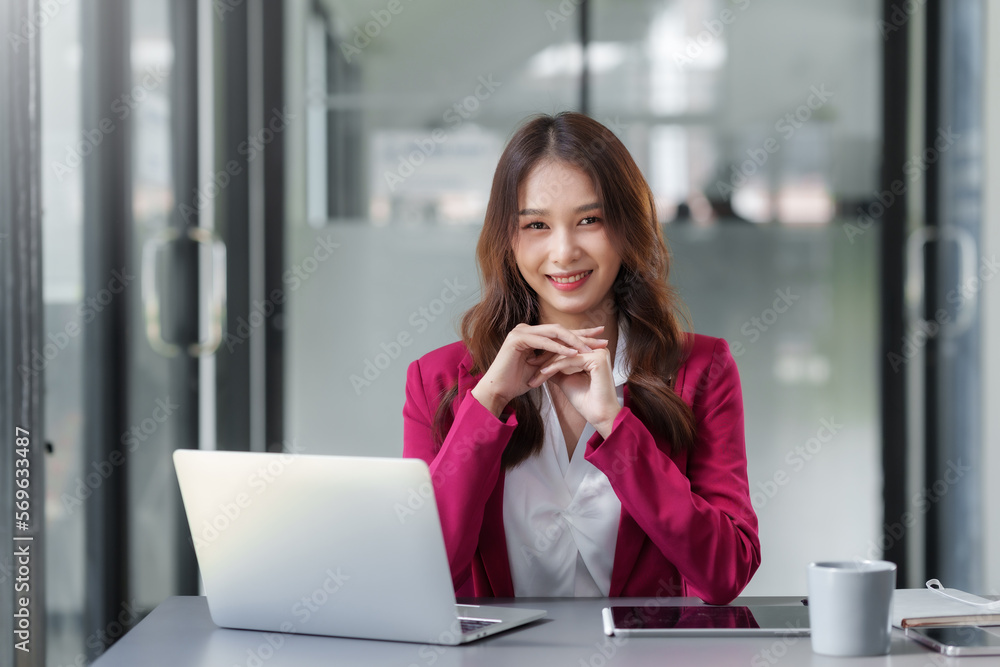 Pretty Asian businesswoman sitting smiling and happy working on laptop computer and document in office. Looking at camera. Pretty Asian businesswoman sitting smiling and happy working on laptop computer and document in office. Looking at camera.
