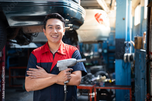 Selective focus of mid-adult Asian male mechanic in red blue uniform, standing hold an impact wrench with arms folded and smiling at camera with blurred garage in background. Copy space on the right.