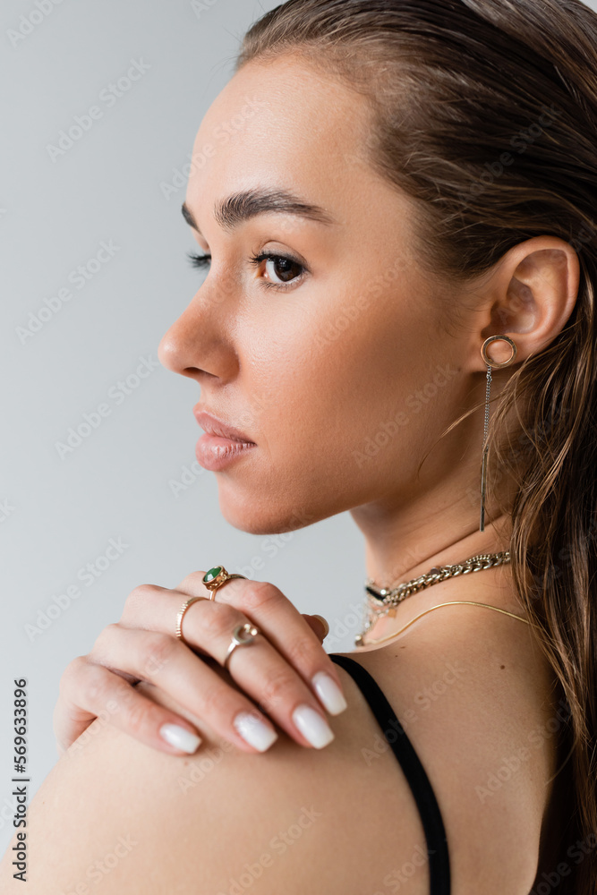 portrait of brunette woman in black top looking away while touching shoulder isolated on grey.