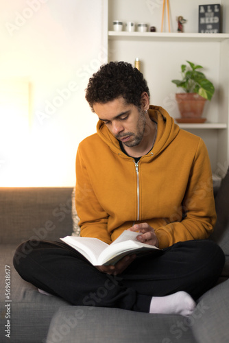 Biracial man reading a book sitting on the sofa at home in casual clothes