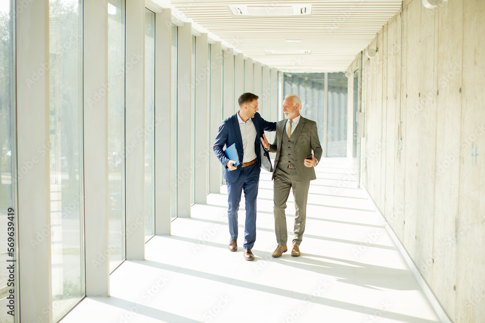 Young and a senior businessman walk down an office hallway, deep in conversation