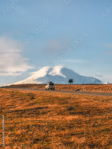 A silver SUV is driving along the highway route to the dawn snow-covered Elbrus. Autumn beautiful road through the mountain desert. Morning mountain valley with a serpentine road. Vertical view.