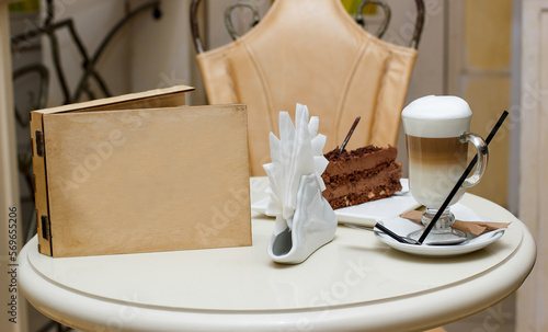 on a white table in a cafe there is a cup of coffee with foam, a saucer with a cake and napkins. Wooden box for the logo. A chair pushed behind a table.