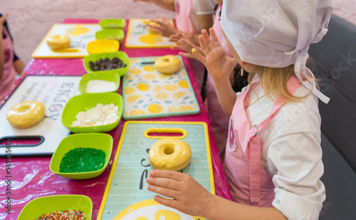 girls at the master class make colored donuts. The ingredients are in boxes. The girl is wearing an apron and cap. girl licking her fingers
