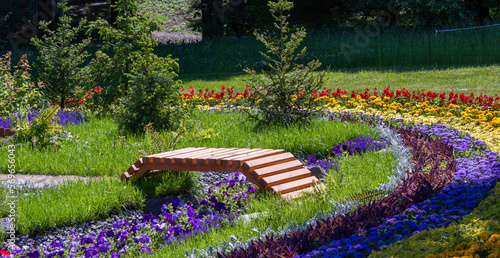Wooden bridge across the moat in the flowerbed. Multicolored flowers. Red, yellow, blue flowers. landscape design