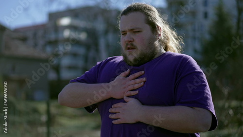 One young overweight man having chest pains standing outdoors. A fat person feeling stressful pain with difficulty in breathing