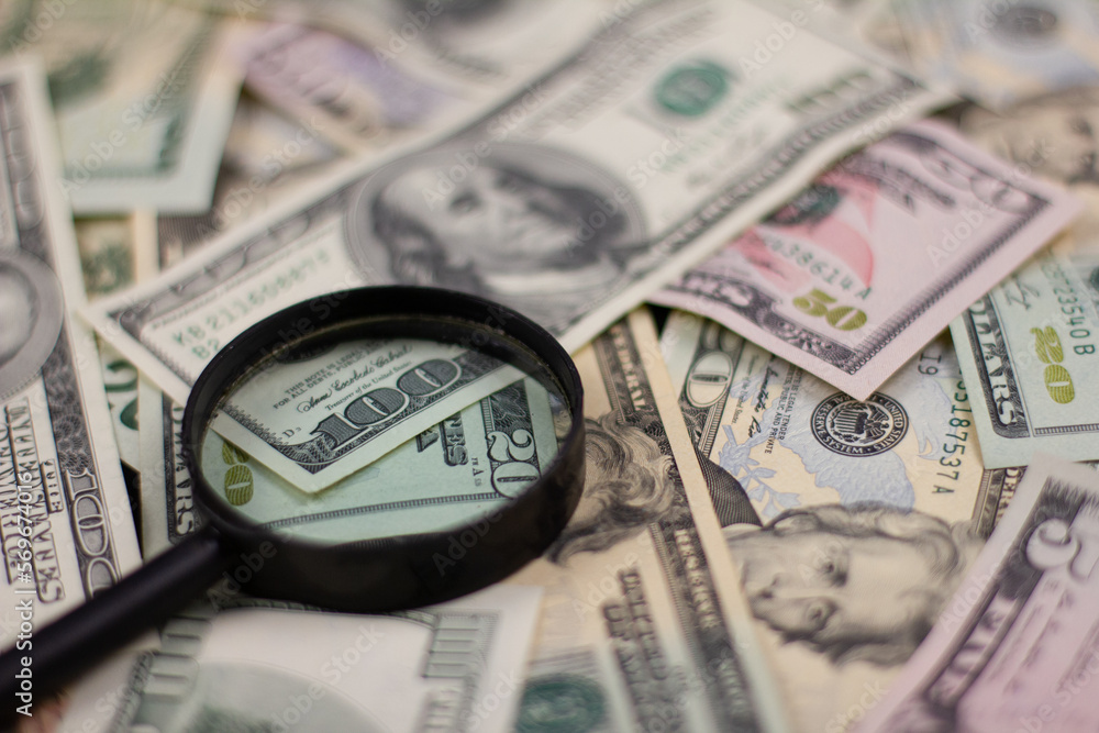 Background from dollar bills and which lies a black magnifying glass Numerous dollar bills are scattered across the table forming a solid background There is a magnifying glass on paper dollar bills. Background from dollar bills and which lies a black magnifying glass Numerous dollar bills are scattered across the table forming a solid background There is a magnifying glass on paper dollar bills.
