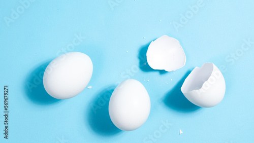Two whole white chicken eggs and one broken on a blue background. The concept of protein nutrition or easter holiday.