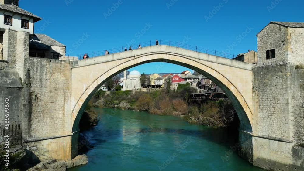 Aerial drone view of the Old Bridge in Mostar city in Bosnia and Herzegovina during sunny day. Blue turquoise colors of Neretva river. Unesco World Heritage Site. People walking over the bridge. Aerial drone view of the Old Bridge in Mostar city in Bosnia and Herzegovina during sunny day. Blue turquoise colors of Neretva river. Unesco World Heritage Site. People walking over the bridge.
