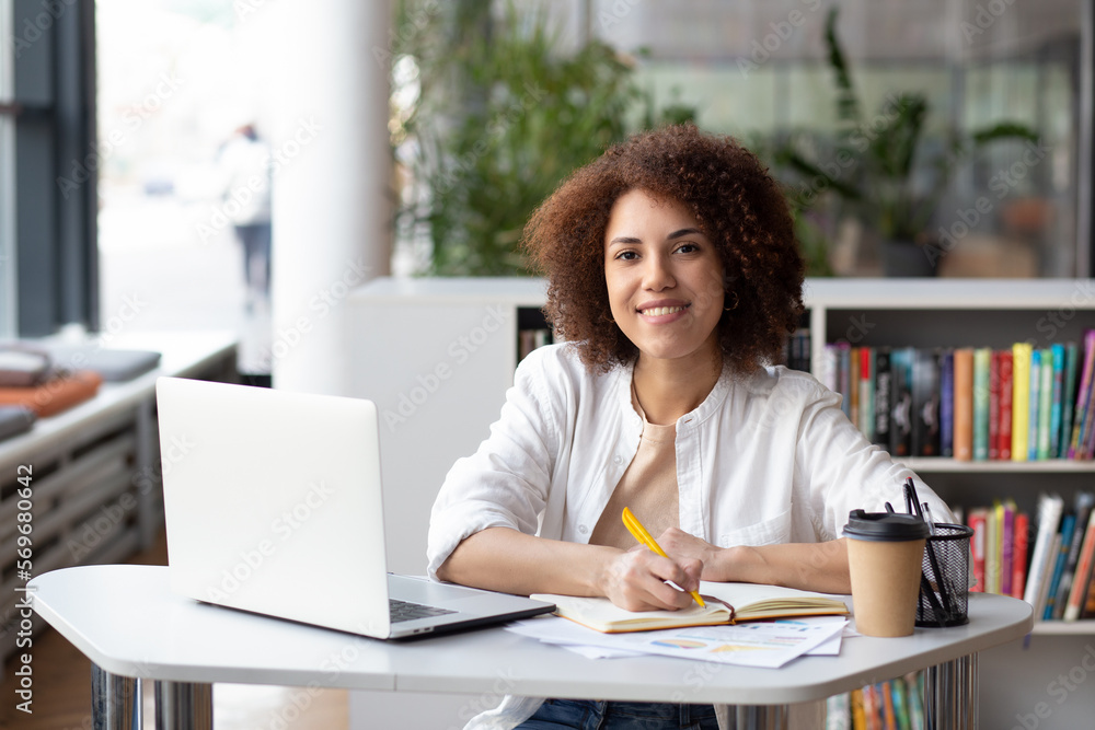 Portrait of a young African American businesswoman or an office worker sitting at a desk with a laptop in a modern office, looking at the camera and smiling Portrait of a young African American businesswoman or an office worker sitting at a desk with a laptop in a modern office, looking at the camera and smiling
