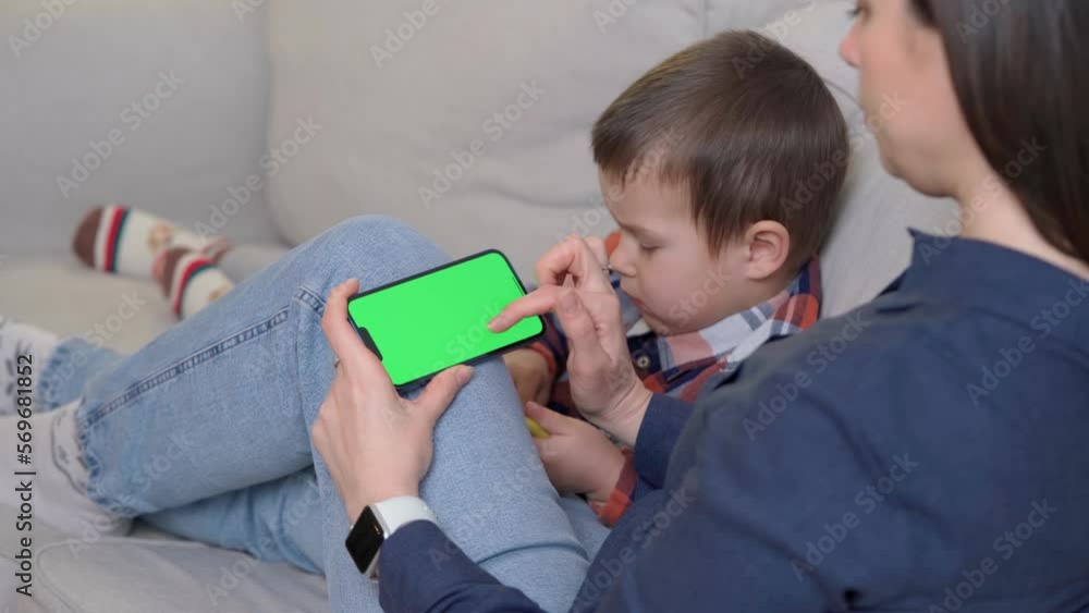 Mother Sits on Sofa with Kid and Using Smartphone with Green Screen, Chroma Key. Boy Plays Mobile Games On Phone. Scrolling Through Social Media or Online Shop Mother Sits on Sofa with Kid and Using Smartphone with Green Screen, Chroma Key. Boy Plays Mobile Games On Phone. Scrolling Through Social Media or Online Shop