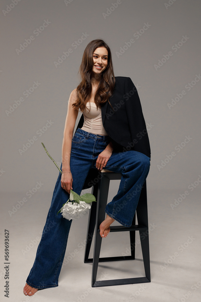 Φωτογραφία A beautiful stylish smiling girl in blue jeans and a black jacket sits on a chair on a gray background and holds white flowers in her hands