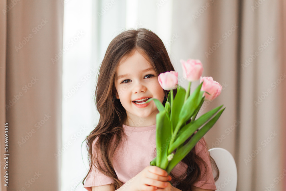 Portrait of a little girl with long dark hair close-up. The baby hugs a bouquet of fresh, delicate pink tulips. A gift for the holiday, spring time. Portrait of a little girl with long dark hair close-up. The baby hugs a bouquet of fresh, delicate pink tulips. A gift for the holiday, spring time.