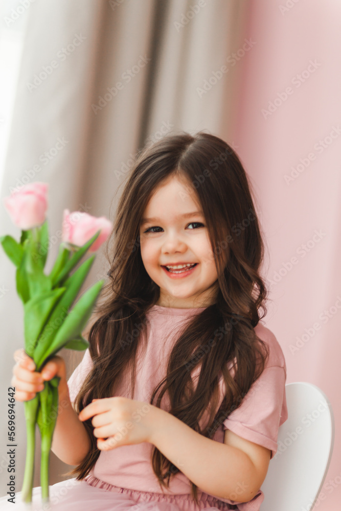 Portrait of a little girl with long dark hair close-up. The baby hugs a bouquet of fresh, delicate pink tulips. A gift for the holiday, spring time. Portrait of a little girl with long dark hair close-up. The baby hugs a bouquet of fresh, delicate pink tulips. A gift for the holiday, spring time.