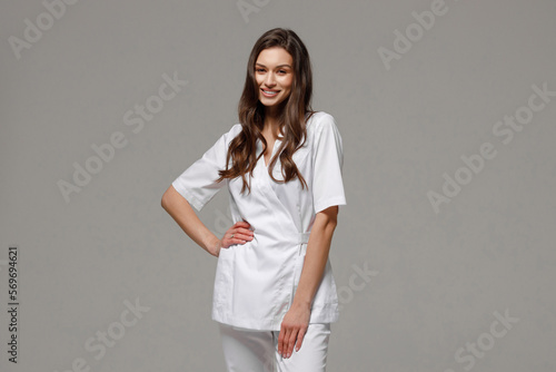 A beautiful young cosmetologist doctor in a white medical suit is smiling at the camera.