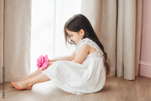 A little girl in a white dress is sitting near the window and holding a rose. A happy child at the window with a flower.