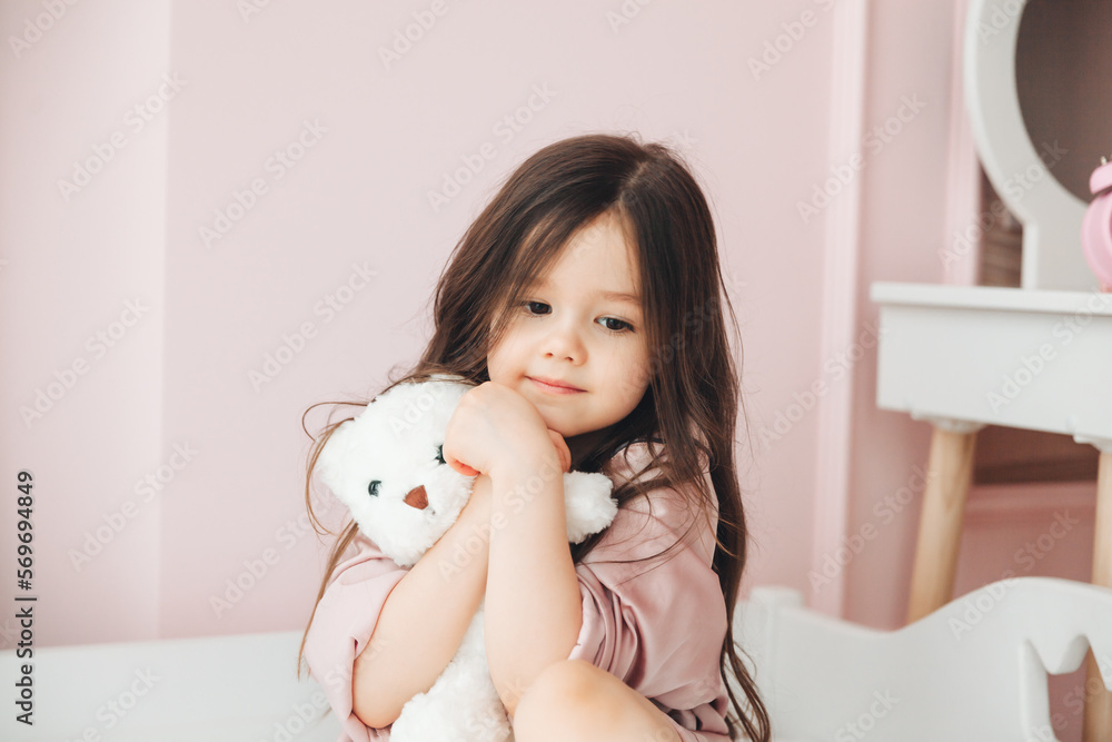 Cute little girl with long hair hugs a teddy bear, looks at the camera and smiles while sitting on her bed at home Cute little girl with long hair hugs a teddy bear, looks at the camera and smiles while sitting on her bed at home