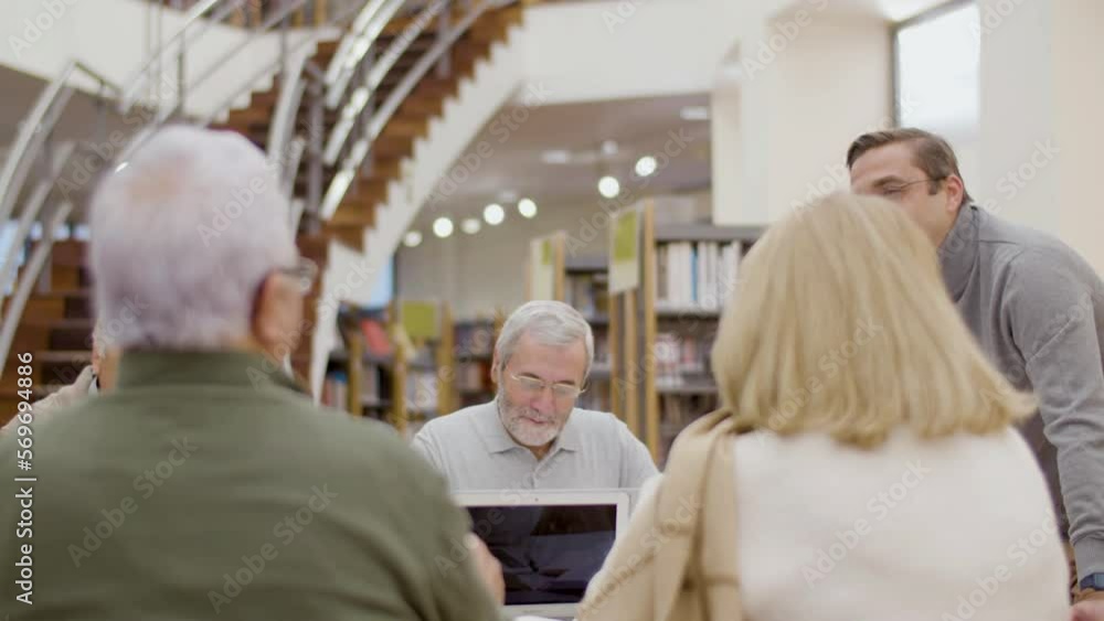 Old people talking with teacher during class in library. Medium shot of man in eyeglasses conducting lesson with senior people. Group of elderly people studying together. Education, retirement concept Old people talking with teacher during class in library. Medium shot of man in eyeglasses conducting lesson with senior people. Group of elderly people studying together. Education, retirement concept