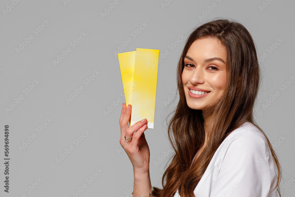 Φωτογραφία A beautiful young cosmetologist doctor in a white medical suit smiles at the camera and holds cosmetology products in her hands