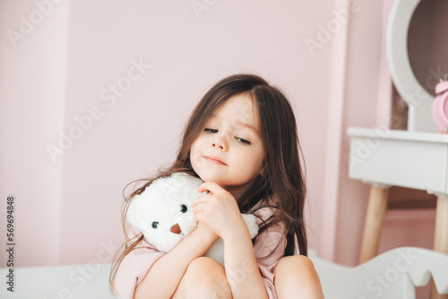 Cute little girl with long hair hugs a teddy bear, looks at the camera and smiles while sitting on her bed at home