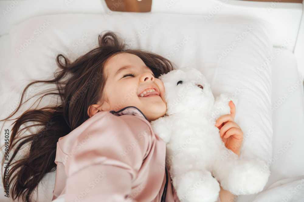 a little dark-haired girl is sleeping in bed, hugging a teddy bear under a white blanket.the child is in bed in the morning. a little dark-haired girl is sleeping in bed, hugging a teddy bear under a white blanket.the child is in bed in the morning.