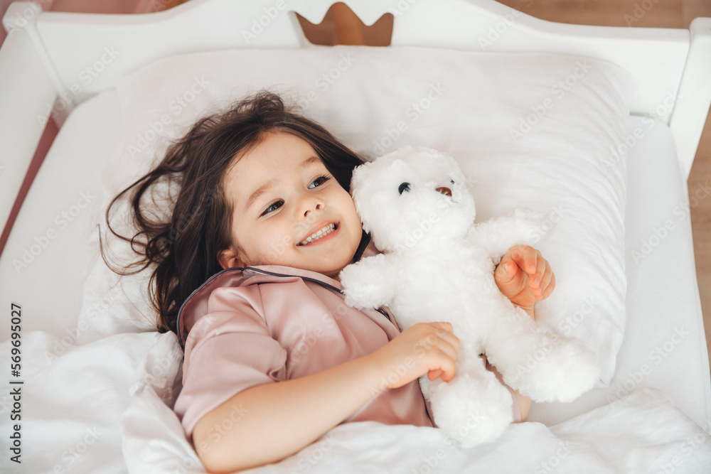 a little dark-haired girl is sleeping in bed, hugging a teddy bear under a white blanket.the child is in bed in the morning. a little dark-haired girl is sleeping in bed, hugging a teddy bear under a white blanket.the child is in bed in the morning.